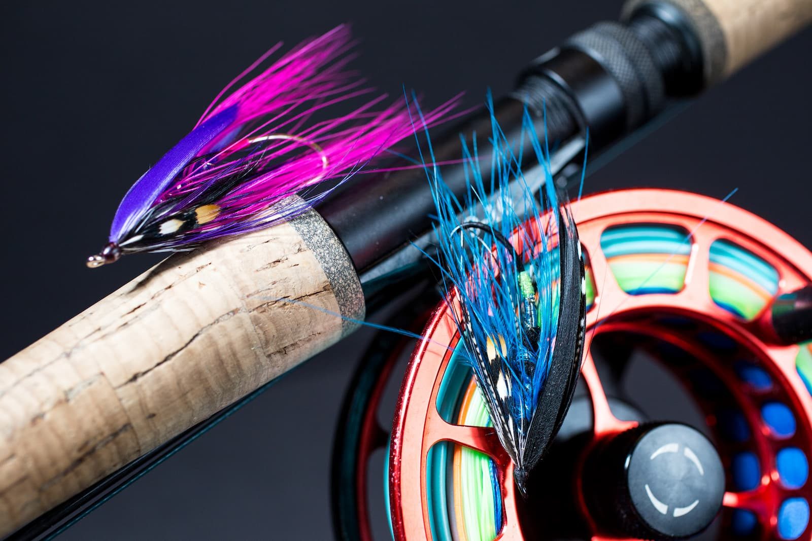 Hand-tied fishing fly in pink and blue on a cork rod handle beside a reel